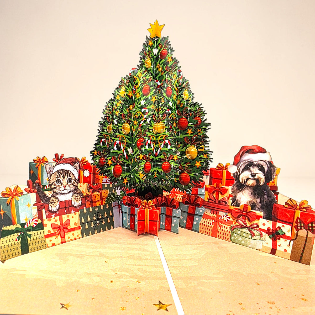 A 3D pop-up cardboard Christmas scene featuring a decorated Christmas tree, surrounded by wrapped presents and cartoon cat and dog characters wearing Santa hats, set against a plain beige background.