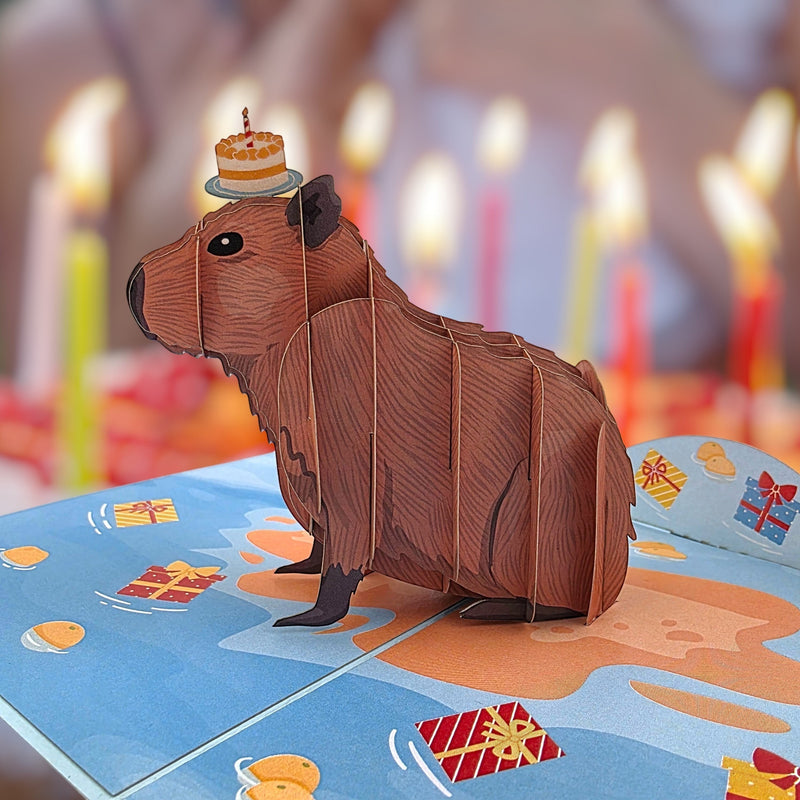 Color Pop Cards 3D pop-up birthday card featuring a brown capybara wearing a small cake on its head, surrounded by festive gifts and confetti.