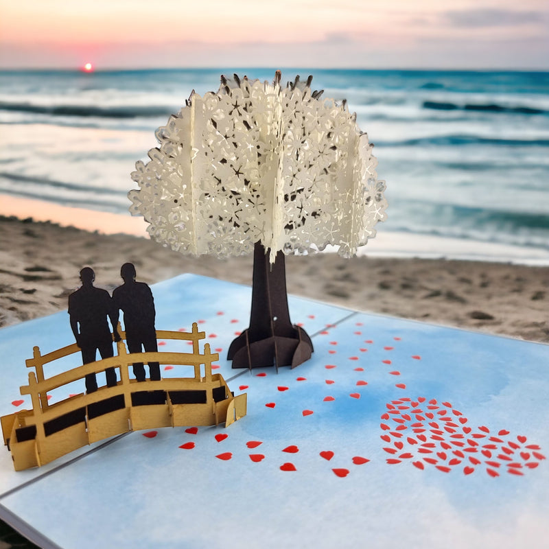 Color Pop Cards pop-up card with a white cherry blossom tree, a bridge, and a same-sex male couple. Red hearts scatter on the beach scene.