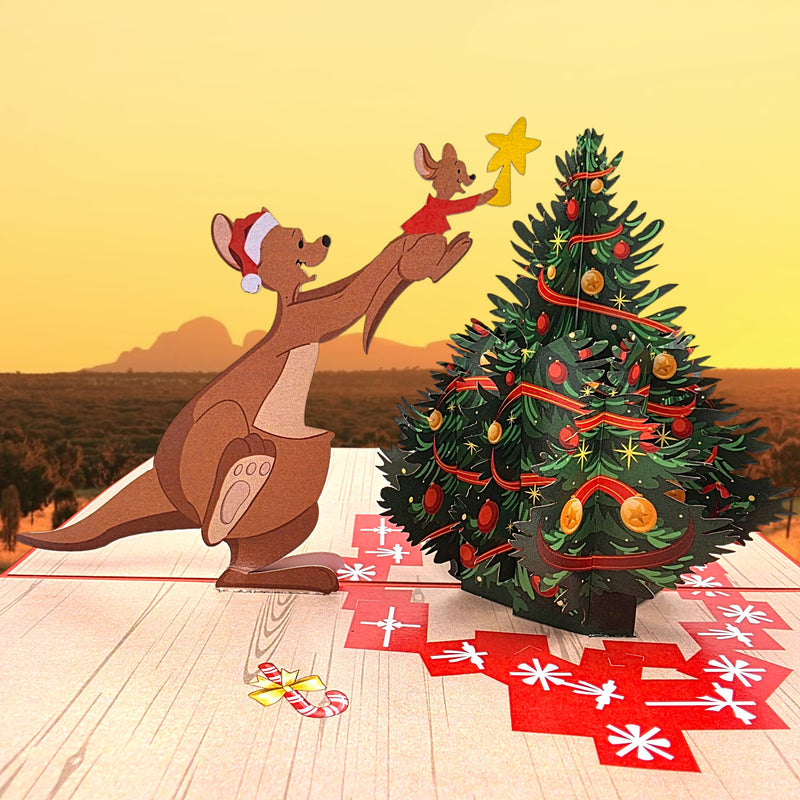 A 3D pop-up cardboard Christmas scene featuring a kangaroo wearing a Santa hat decorating a Christmas tree with a mouse and a star, set against a warm sunset landscape background.