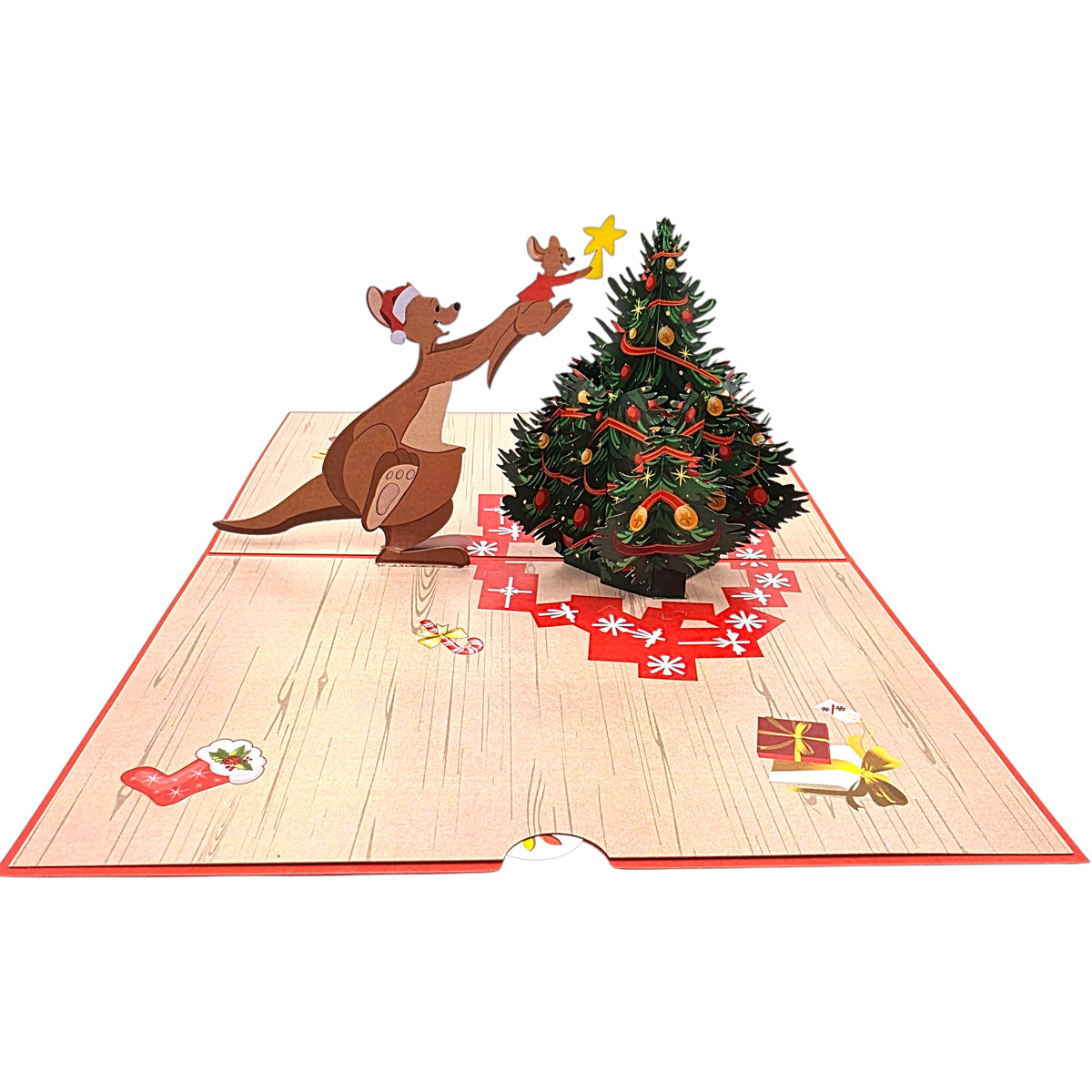 A 3D pop-up cardboard Christmas gift box featuring a kangaroo decorating a Christmas tree on a wooden floor base with red gift and stocking details, shown against a plain white background.